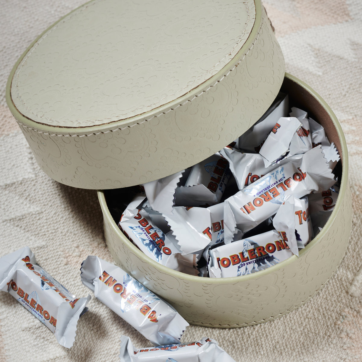 Dose rund flach ICON aus Leder geprägt im farbton foggy, gefüllt mit toblerone schokolade